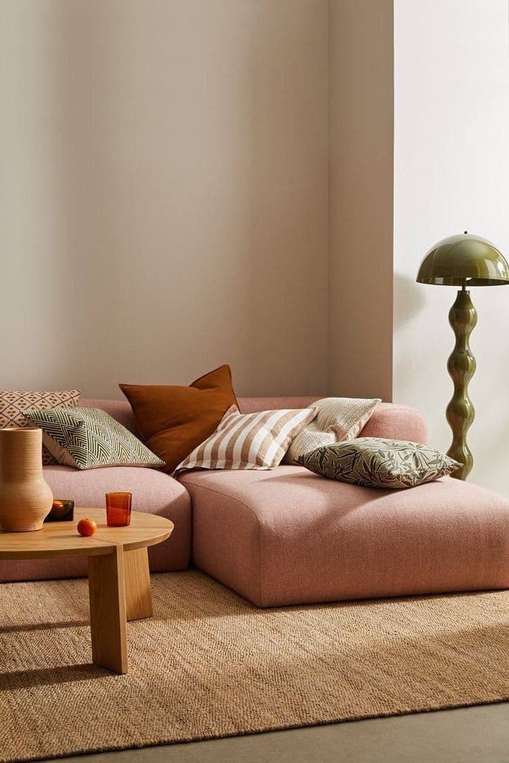 En låg modulsoffa i rosenrosa med kuddar i rost, ränder och grönt, ett skulpterat soffbord i ek, en jutematta och en olivgrön golvlampa med svarvad fot.