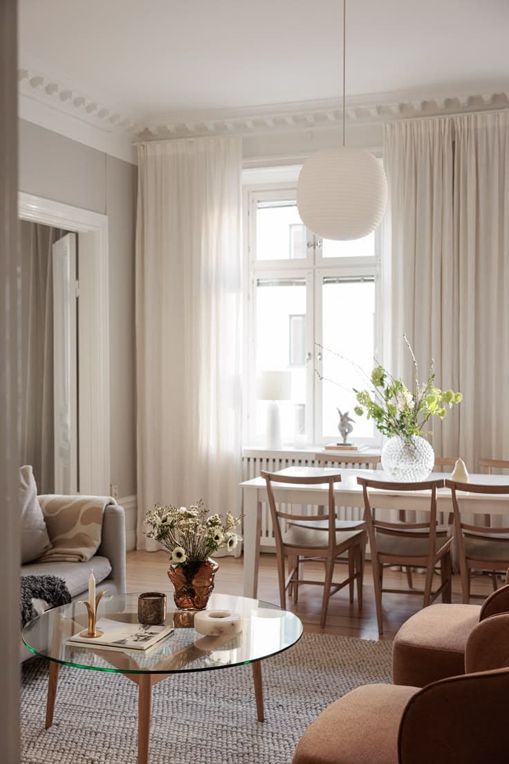 Ett runt glasbord på ekben, rostfärgade bouclé-fåtöljer och ett matbord i ek med pinnstolar står under en klotlampa mellan golvlånga tunna gardiner.