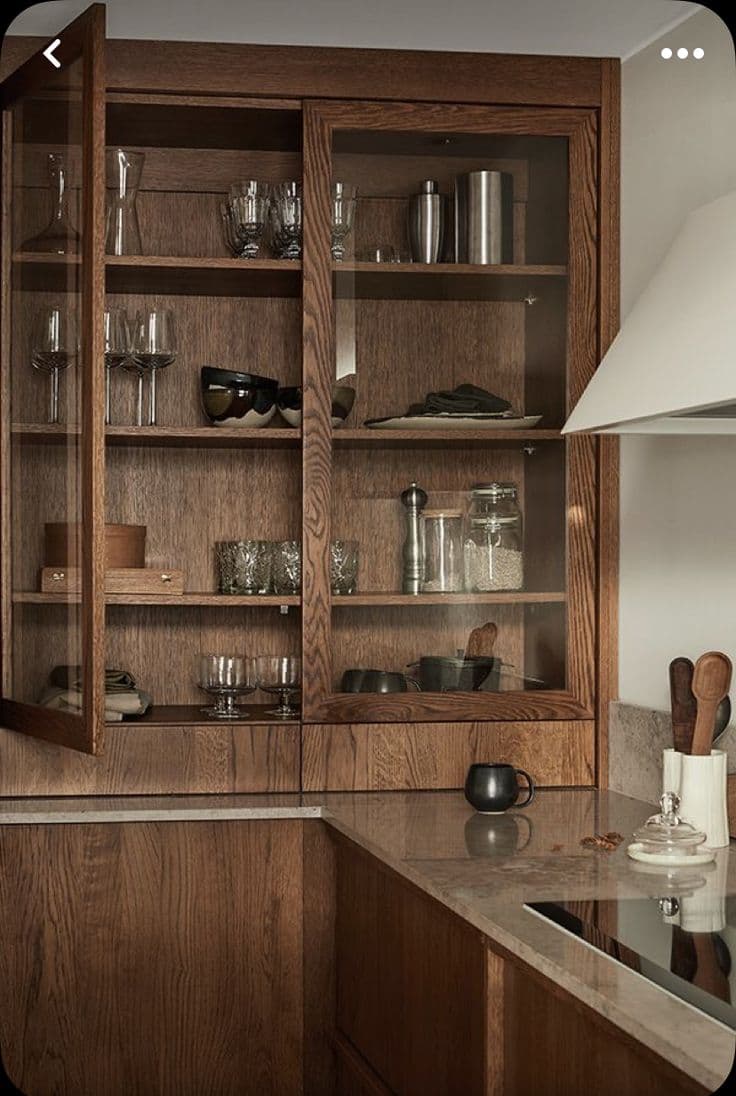 Ett kök med mörkbetsade ekskåp med glasdörrar fyllda med glas, matchande underskåp i ek, en beige stenbänk och en vit köksfläkt.