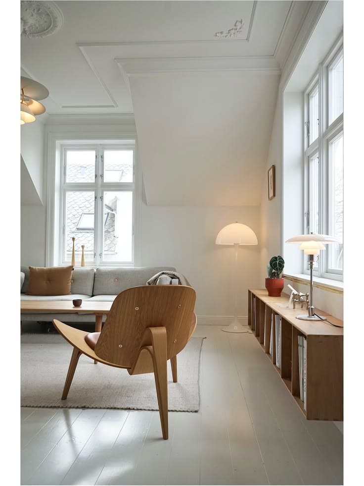 Ett vitmålat vindsrum med gråvita golvbrädor, en skalstol i formpressad ek, en låg ekhylla, en svamplampa på golvet och en grå soffa under stuckatur i taket.