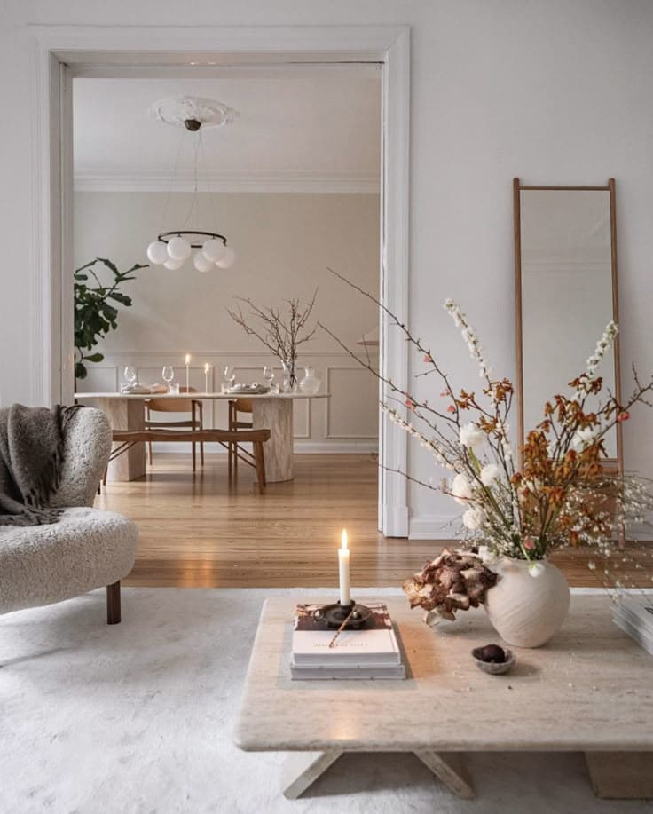 Ett soffbord i travertin med blommande grenar och ett ljus står på en vit ullmatta bredvid en bouclé-fåtölj, med utsikt in i en matsal med klotkrona.