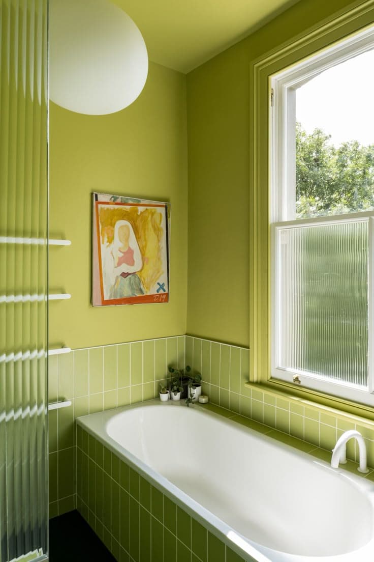Ett limegrönt badrum med grönt stående kakel, räfflat glas, en stor glob-lampa och en inramad målning.