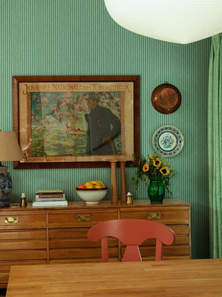 Grönrandig tapet bildar fond åt en teakbyrå med mässingsbeslag, en inramad vintageaffisch, en korallröd målad stol och ett matbord i furu.