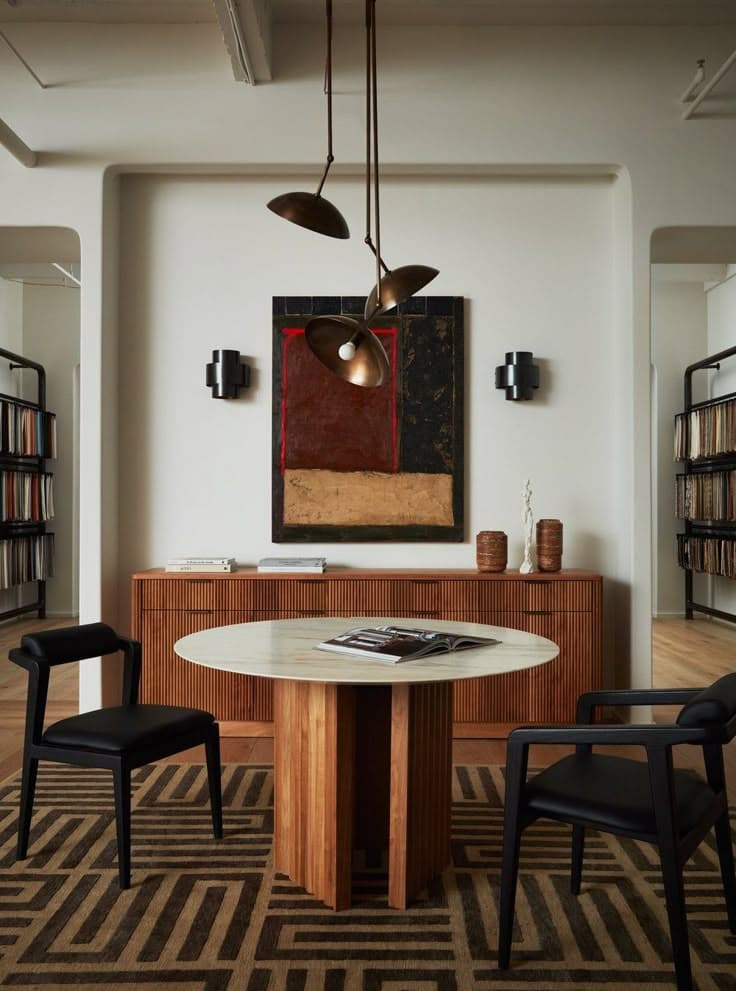 Ett pelarbord i valnöt med ljus stenskiva, svarta stolar, en sideboard i valnöt, bronsfärgade taklampor och en brun geometrisk matta mot vita putsväggar.