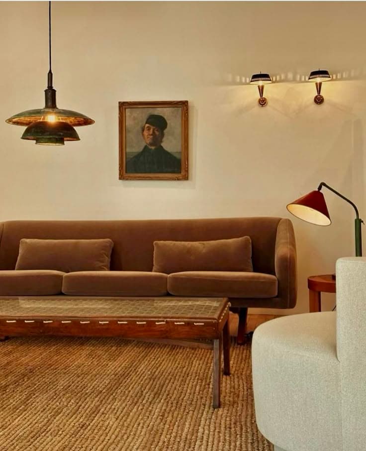 En gråbrun sammetssoffa under en PH-lampa i mässing, ett gammalt porträtt, en röd läslampa och en jutematta.