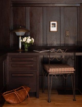 Mörkbetsad ekpanel omsluter ett inbyggt skrivbord med lådor, en smidesstol med randig dyna, en svamplampa och en flätad läderkorg.