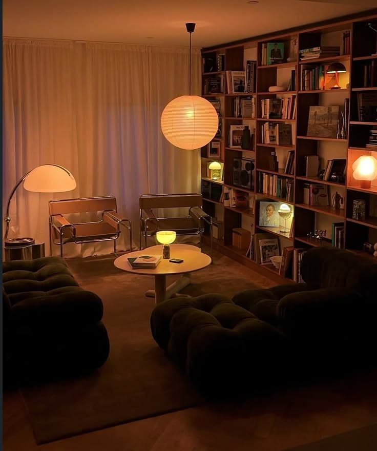 Ett bibliotekslikt vardagsrum med bokhyllor i valnöt från golv till tak, en rund rispapperslampa, två Wassilystolar i krom och ljusbrunt läder och djupa olivgröna modulsoffor kring ett runt vitt bord.