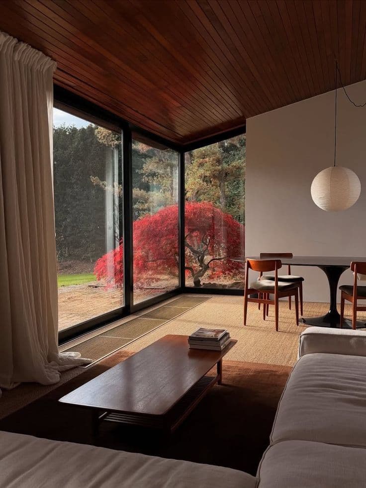 Ett hörn av glas ramar in en röd japansk lönn under ett mörkt rödbrunt trätak, med teaksoffbord, vit soffa, rispapperslampa och vitt tulpanbord med teakstolar.