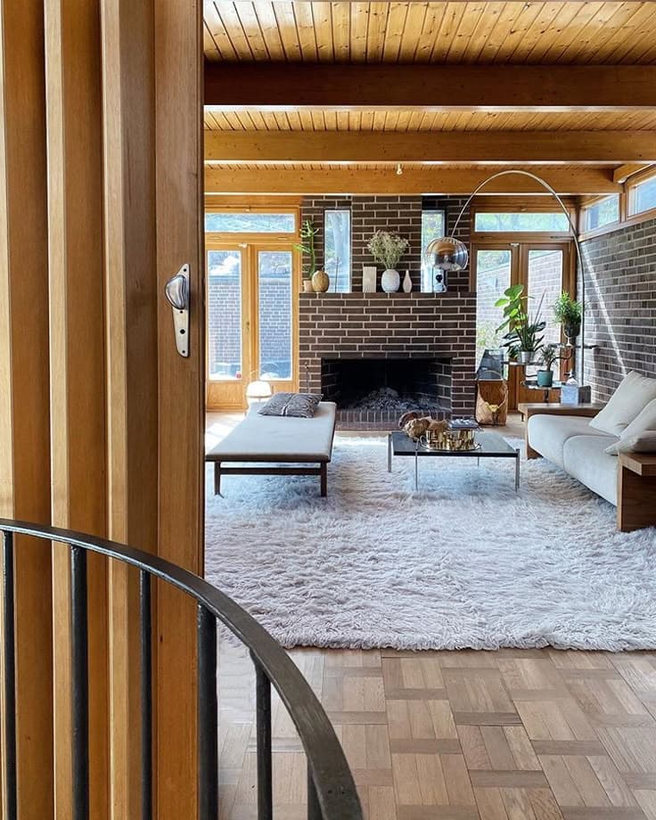 Ett vardagsrum från 60-talet med furupanel i taket, en mörk tegelvägg med öppen spis, en vit ryamatta, en vit dagbädd i Barcelonastil, ett glasbord och parkett i teakton.